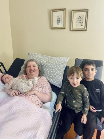 Carm, in a pink pajama top, lying in bed holding her grandson, with her other grandson and grandnephew standing beside her.